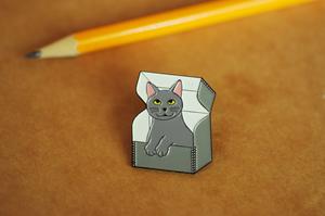 Enamel pin with smiling gray kitty sitting in an archival box with the lid open.